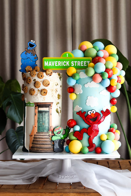 Two-part character themed birthday cake with a street scene and a bright balloon garland