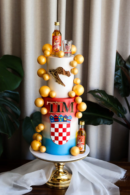 Blue and white celebration cake with Croatian crest detail and a gold balloon garland