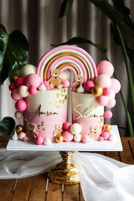 Split pink and white twin birthday cake with a fondant rainbow, teddy bears and balloon garland