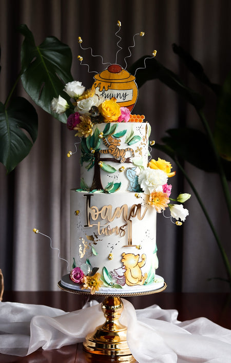 Two-tier storybook themed first birthday cake with a honey pot topper and hand-painted woodland scene