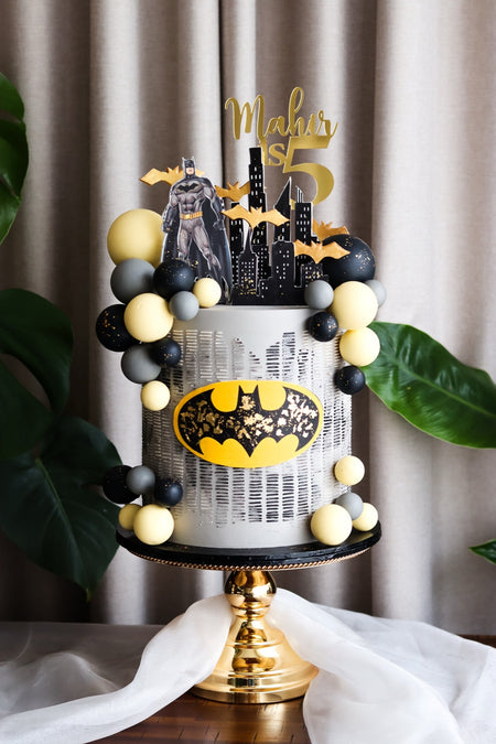 Black and yellow superhero themed birthday cake with a city skyline silhouette and balloon garland