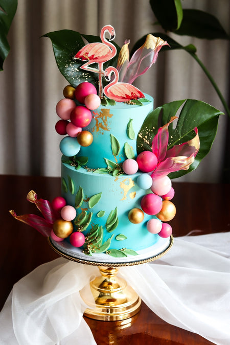 Two-tier teal tropical cake with a flamingo topper, palm leaves and balloon garland