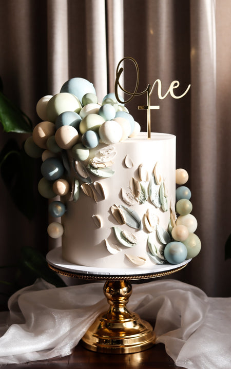 Neutral toned christening cake with sugar petals, a balloon garland and gold cross topper