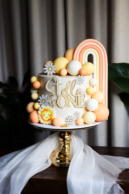 Neutral first birthday cake with a fondant rainbow, daisies and a sunshine topper
