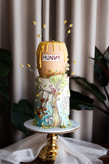 Two-tier storybook cake with a honey pot topper, sugar bees and a hand-painted woodland scene