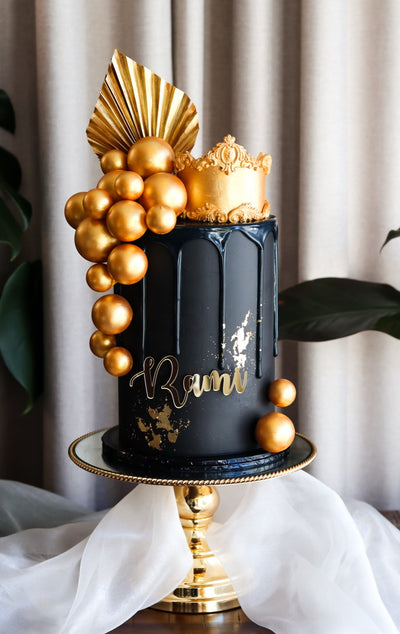 Single tier black drip cake with gold spheres, gold leaf and a fondant crown topper